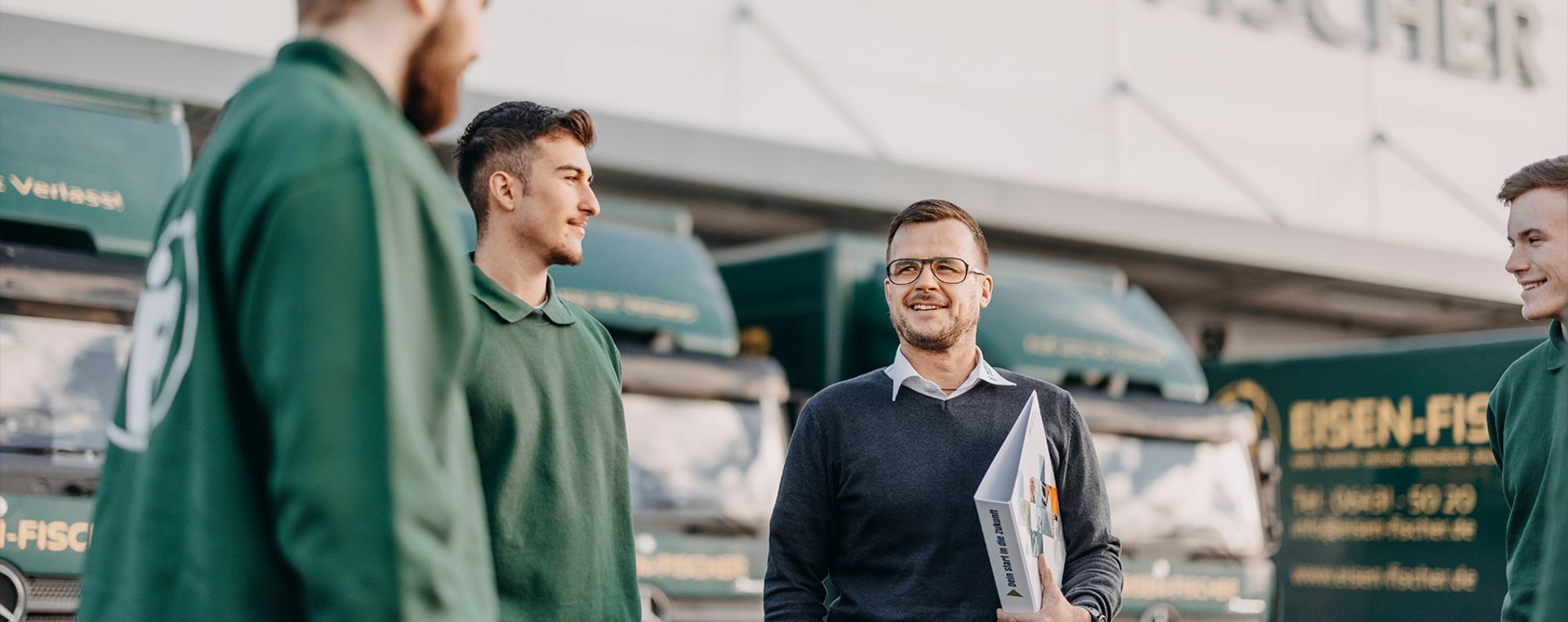 Ein Ausbilder spricht mit Auszubildenden vor Eisen-Fischer-Fahrzeugen. Alle tragen grüne Arbeitskleidung.