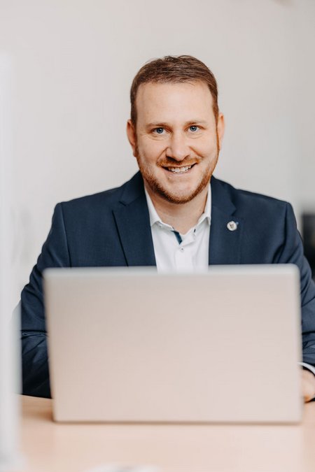 Ein Mann im dunklen Sakko sitzt an einem Schreibtisch vor einem Laptop und lächelt in die Kamera. Der Hintergrund ist neutral und leicht unscharf.