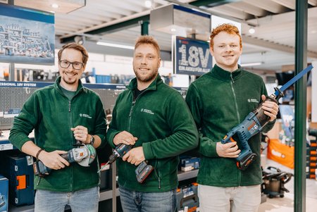 Drei Mitarbeiter in grünen Eisen-Fischer-Fleecejacken stehen in einer Werkzeugabteilung und halten Elektrowerkzeuge in den Händen. Alle drei lächeln in die Kamera.