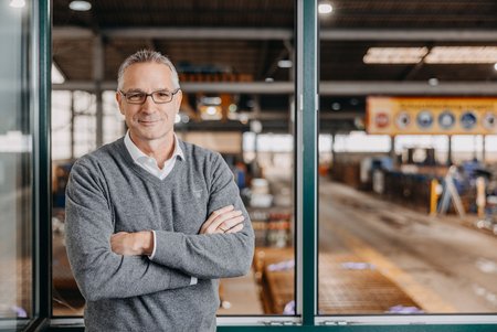 Ein Mann mit Brille und grauem Pullover steht mit verschränkten Armen vor einem großen Hallenfenster. Dahinter ist der Stahlbereich des Unternehmens sichtbar.
