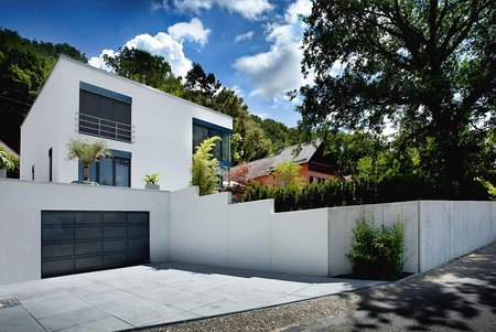 Eckiges, modernes Wohnhaus mit weißen Fassadenflächen, großen Fenstern und dunkelgrauem Garagentor, umgeben von Bäumen.