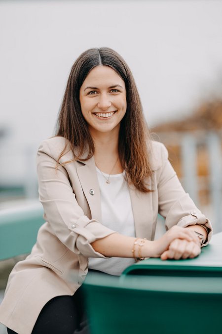 Eine junge Frau mit langen dunklen Haaren sitzt lächelnd an einem Tisch im Freien. Sie trägt einen beigefarbenen Blazer und ein weißes Shirt. Der Hintergrund ist leicht unscharf.