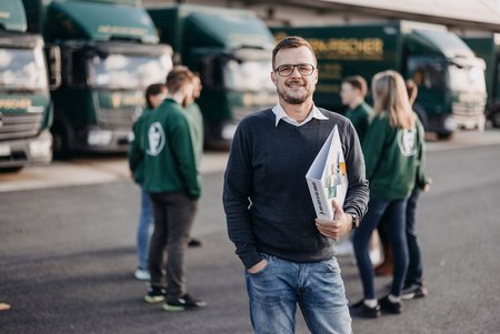 Ein Mitarbeiter steht mit einer Mappe in der Hand vor mehreren grünen Eisen-Fischer-LKWs. Im Hintergrund stehen weitere Mitarbeitende in Eisen-Fischer-Jacken zusammen.