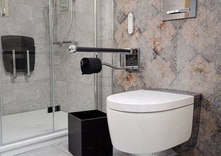 Wandhängendes WC mit ausklappbarem Haltegriff in Chrom, gemusterter Wandfliese in Beige-Blau, daneben eine Glasdusche mit grauen Fliesen und Duschsitz.