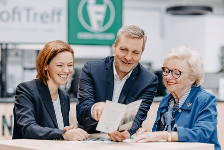 Zwei Frauen und ein Mann sitzen gemeinsam an einem Tisch und schauen lächelnd in eine Broschüre. Im Hintergrund ist ein Logo von Eisen-Fischer zu sehen.