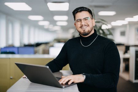 Ein junger Mann mit Brille und schwarzem Rollkragenpullover sitzt an einem Stehtisch im Büro und arbeitet an einem Laptop. Er lächelt in die Kamera, im Hintergrund ist ein Großraumbüro erkennbar.