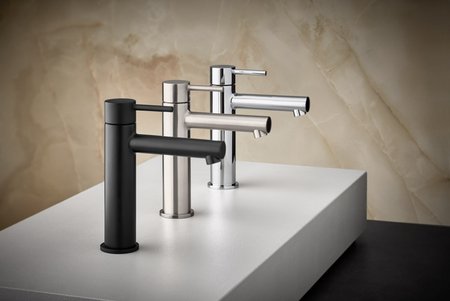 Drei moderne Waschtischarmaturen in Schwarz, Edelstahl und Chrom stehen auf einer weißen Podestfläche vor einer beige-marmorierten Wand.