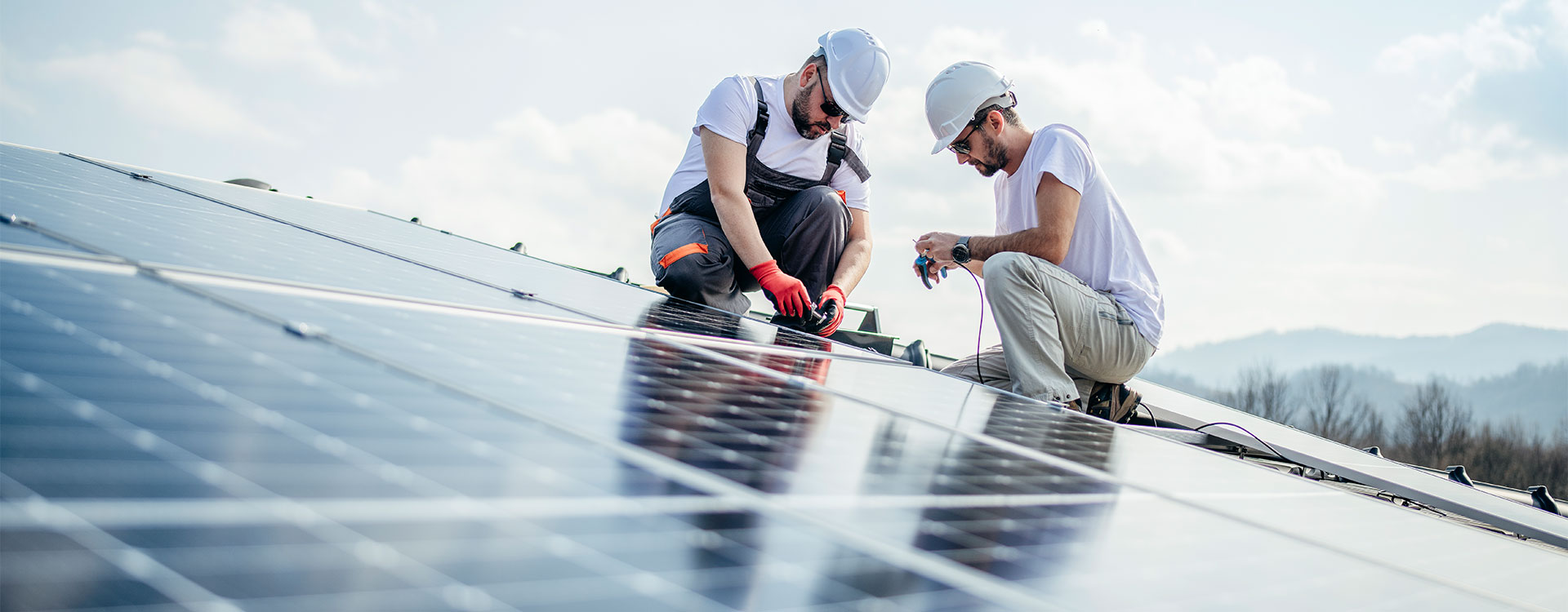 Zwei Techniker installieren Photovoltaikmodule auf einem Dach, tragen Schutzhelme und arbeiten über den Solarpaneelen.