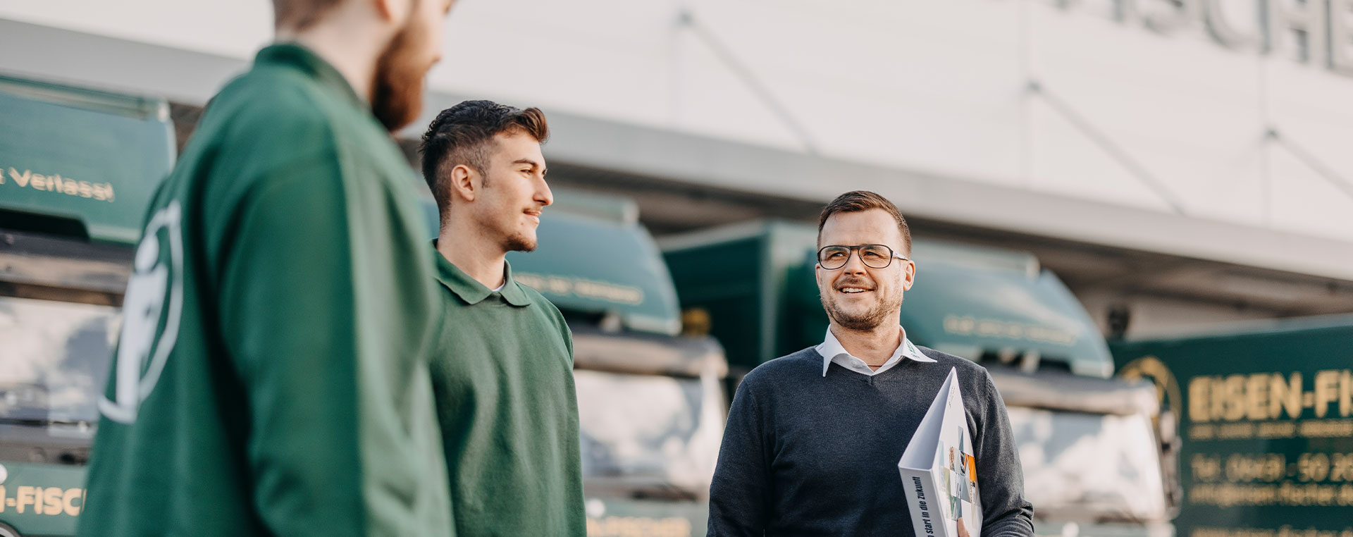Ein Ausbilder spricht mit jungen Mitarbeitenden vor grünen Eisen-Fischer-LKWs. Alle tragen grüne Arbeitskleidung.
