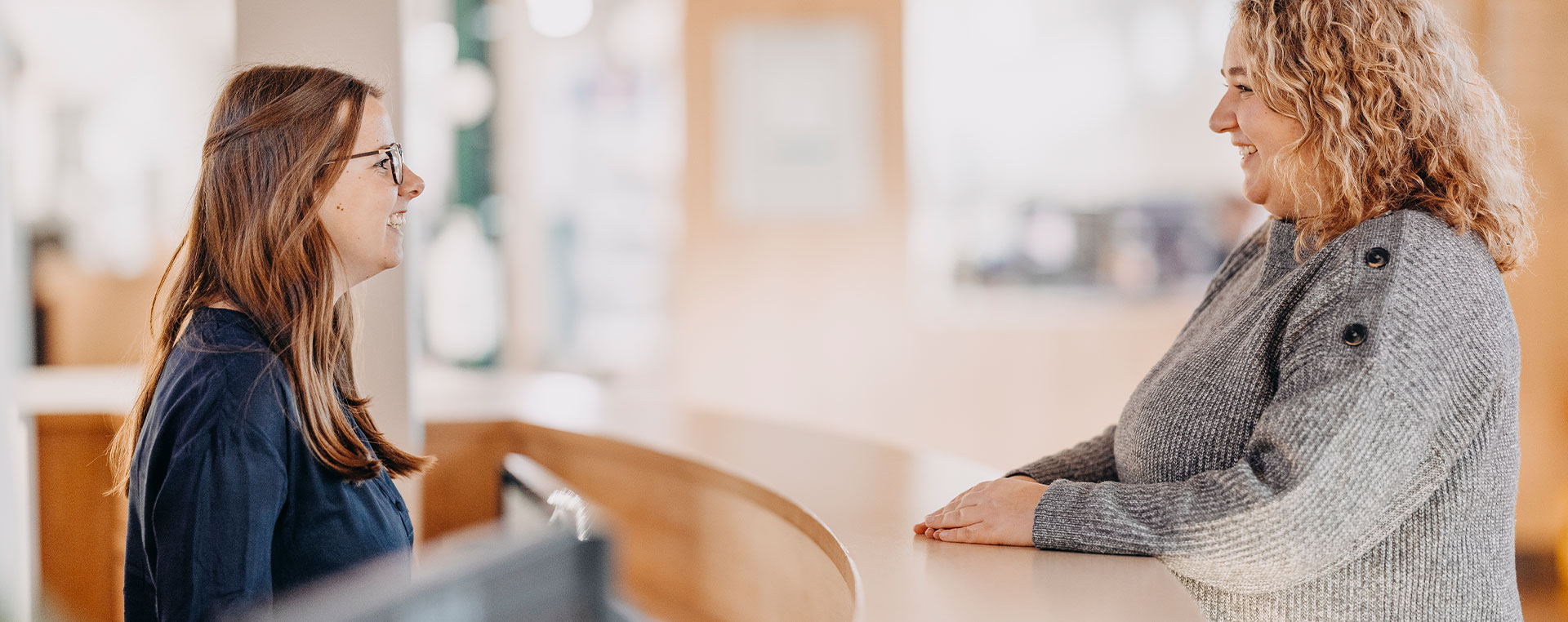 Zwei Mitarbeiterinnen unterhalten sich freundlich an einem Empfangstresen in einem hellen Innenraum.