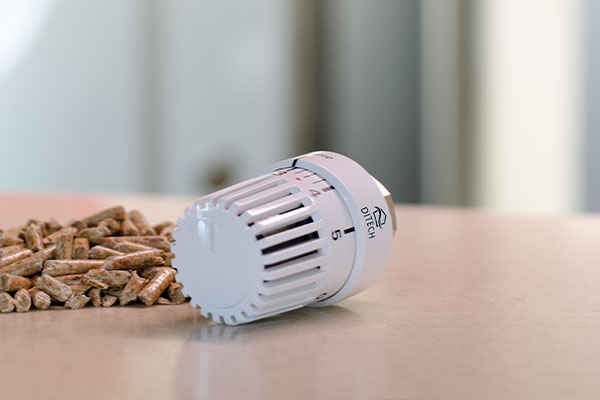 Weißer Thermostatkopf liegt auf einer Tischplatte neben kleinen Holzpellets, im Hintergrund verschwommene Wohnumgebung.