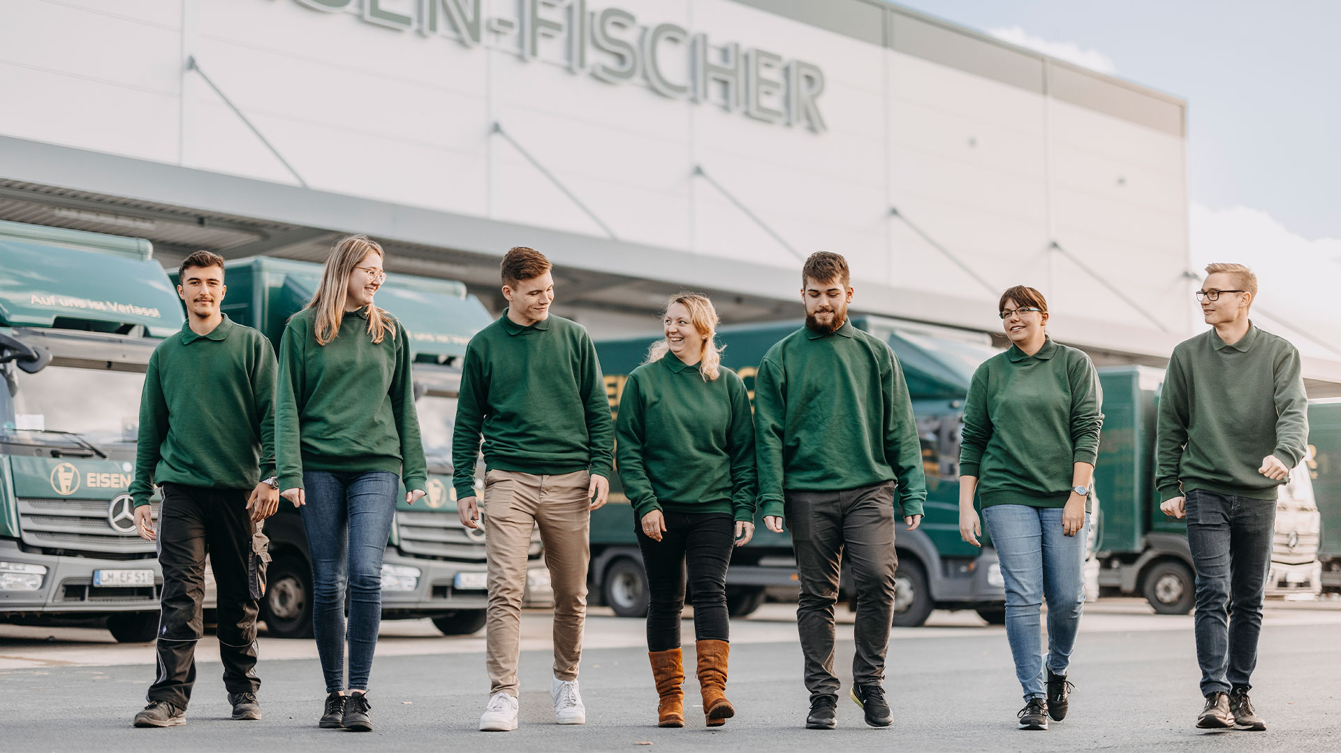 Gruppe junger Mitarbeitender im grünen Eisen-Fischer-Outfit läuft gemeinsam vor dem Unternehmensgebäude.